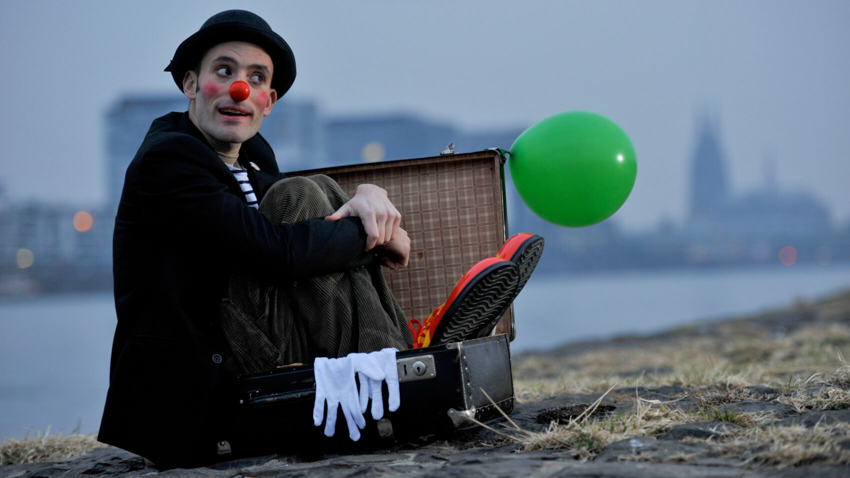 Clown Olli sitzt im Koffer mit roter Nase und riesigen roten Schuhen