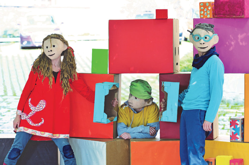Drei Schauspieler*innen mit Masken über den Köpfen, die ein Mädchen und zwei Jungen darstellen. Sie stehen bei einer Mauer aus bunten Würfeln.